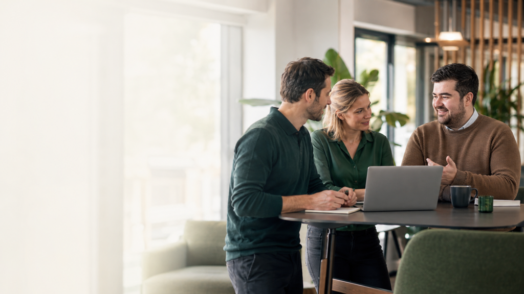 Drie collega's overleggen aan een statafel met een laptop in een licht kantoor.